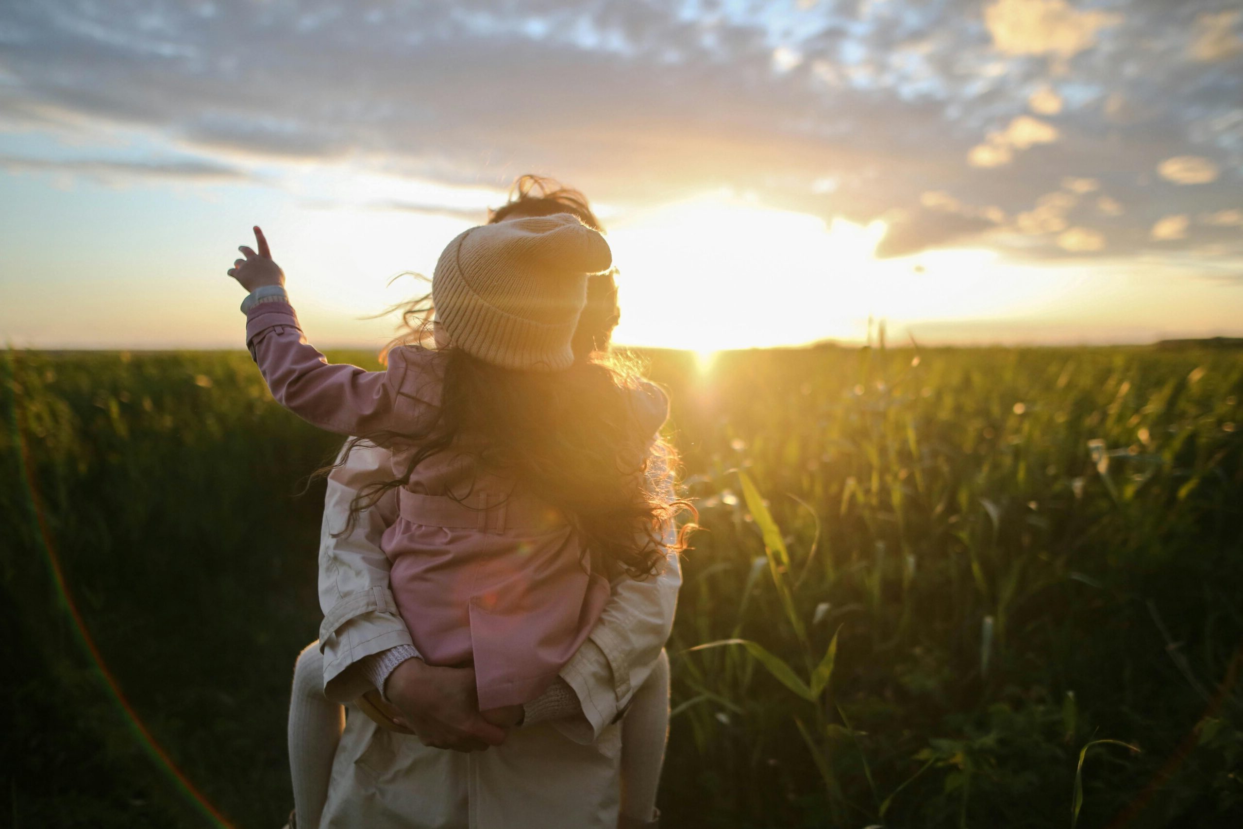 A mother and daughter embrace and point at the sunset in a grassy field.